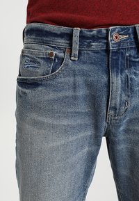 Mid-blå denimjeans med synliga framfickor, bältesöglor och kopparfärgade nitar, matchade med en röd överdel.