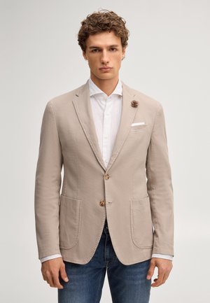 Młody mężczyzna w beżowej marynarce z dwoma guzikami, białej koszuli i niebieskich dżinsach, stojący na tle jednolitego jasnego tła.