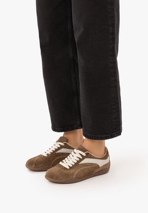 Persoon draagt bruine suède sneakers met witte strepen en witte veters, gecombineerd met zwarte cropped broek.
