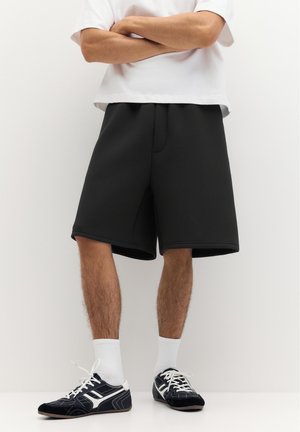 Homme portant un short noir au genou, des chaussettes blanches, des baskets noires et blanches et un T-shirt blanc sur un fond clair uni.