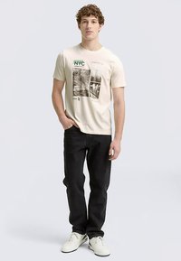 Beige värinen graafinen t-paita, jossa NYC-kuvio, lyhyet hihat, mustat farkut ja valkoiset lenkkarit, seisoo vaaleaa taustaa vasten.