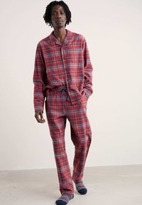 Punavihrine ruudullinen pyjama-asusetti, pitkähihainen nappipaita ja naruilla varustetut housut, valmistettu pehmeästä kankaasta. Mallilla on jalassaan värikkäät kuviolliset sukat.