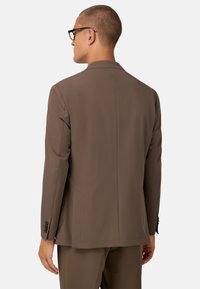 Ruskea puku takki, jossa on muotoiltu istuvuus, takana on yksi halkio ja nappikoristeet hihassa. Sujuva kangasperhe.