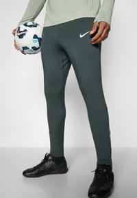 Grønne sportsleggings med en tætsiddende pasform, der har et hvidt Nike-logo og side-syninger. Sort fodbold holdt i hånden.
