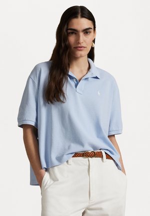 Lyseblå poloskjorte laget av teksturert stoff, med en tre-knapps placket og brodert logo. Båret med hvite bukser og et flettet belte.