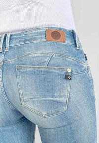 Vaqueros de mezclilla azul claro con una textura suave, que tienen un parche de cuero marrón en la cintura y una etiqueta negra en el bolsillo trasero.