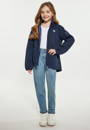 Mørkeblå zip-up jakke med struktureret overflade, der har høj krave og logo på brystet, parret med lyseblå jeans og hvide sneakers.