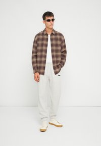 Brun og beige rutete flanellskjorte, hvit t-skjorte, lys grå joggebukser og beige joggesko. Tilbehør inkluderer brune solbriller.