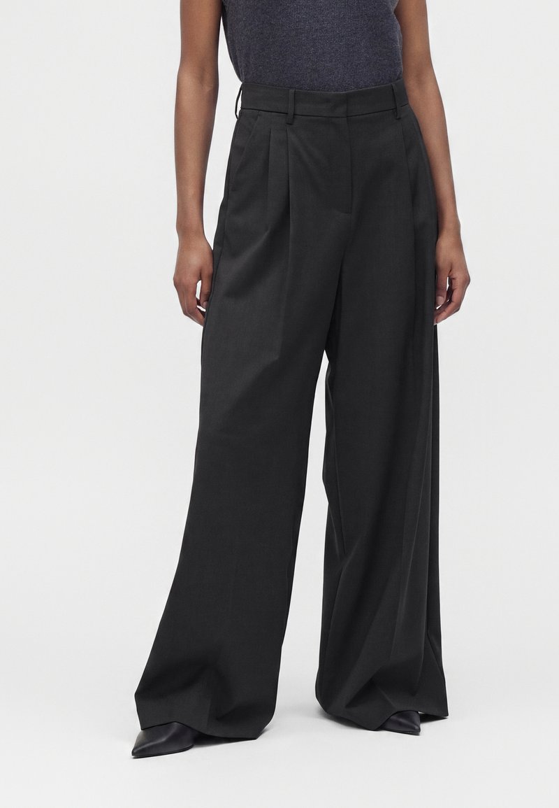 Personne portant un pantalon large noir taille haute avec plis, associé à des chaussures pointues noires et un haut sans manches gris foncé.