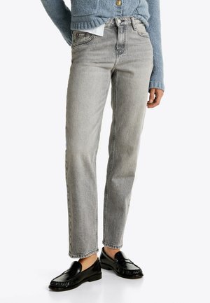 Osoba nosząca szare jeansy o prostych nogawkach, czarne mokasyny i niebieski dziergany sweter ze złotymi guzikami, stojąca na jasnym tle.