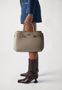 Mujer con botas de cuero marrón y falda de mezclilla azul sosteniendo un bolso taupe de Guess con herrajes dorados a su lado.