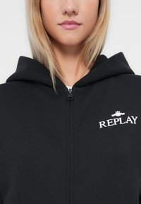 Čierna mikina s kapucňou na zips vyrobená z jemného materiálu, s logom "REPLAY" v bielej farbe na ľavej strane hrude a podšívkou v kapucni.