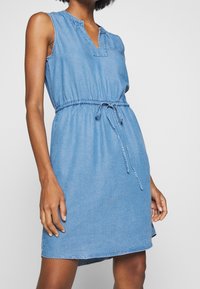 Denim-mekko ilman hihoja, jossa on V-pääntie, joustava kuminauha vyötäröllä ja solmiyksityiskohta. Vaaleansininen väri, sileä tekstuuri ja polvimittainen helma.