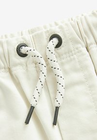 Beige shorts med elastisk talje og snøre. Snøret er hvidt med sorte detaljer og fastgjort med metaløjer.