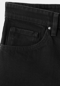 Gros plan sur un jean en denim noir montrant la poche avant, la poche à monnaie, la boucle de ceinture et le détail des rivets métalliques.