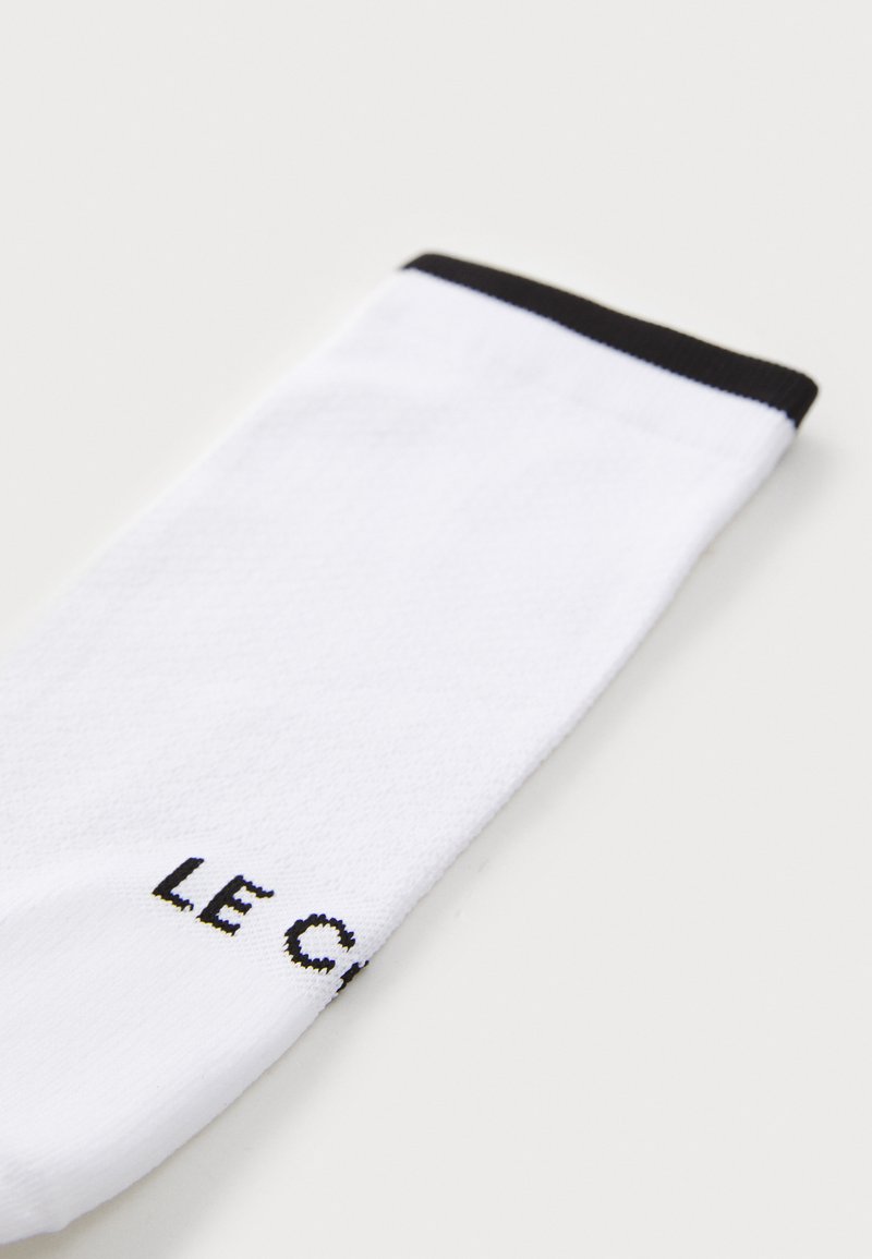 Weißer Socken mit strukturiertem Oberflächenmaterial und einem schwarzen gerippten Bündchen. Der Markenname "LE C" ist in fettem schwarzen Text nahe dem Bündchen aufgedruckt.