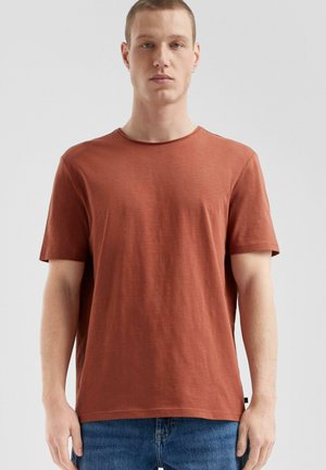 T-shirt w kolorze rdzy z bawełny, z okrągłym dekoltem, krótkimi rękawami i luźnym krojem. Gładka tekstura z minimalnymi wzorami i detalem w postaci metki z boku.