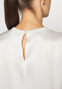 Blouse en soie beige clair avec un col rond, dotée d'une fente à l'arrière fermée par un petit bouton et d'un col texturé.