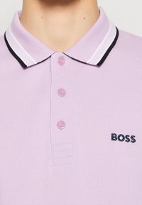 Gros plan sur un polo violet clair avec un col rayé blanc et noir, trois boutons, et « BOSS » brodé sur la poitrine.