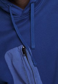 Felpa blu con una tasca con zip di un blu più chiaro in contrasto, stringhe di chiusura e una texture di tessuto morbido. Logo ricamato sulla tasca.