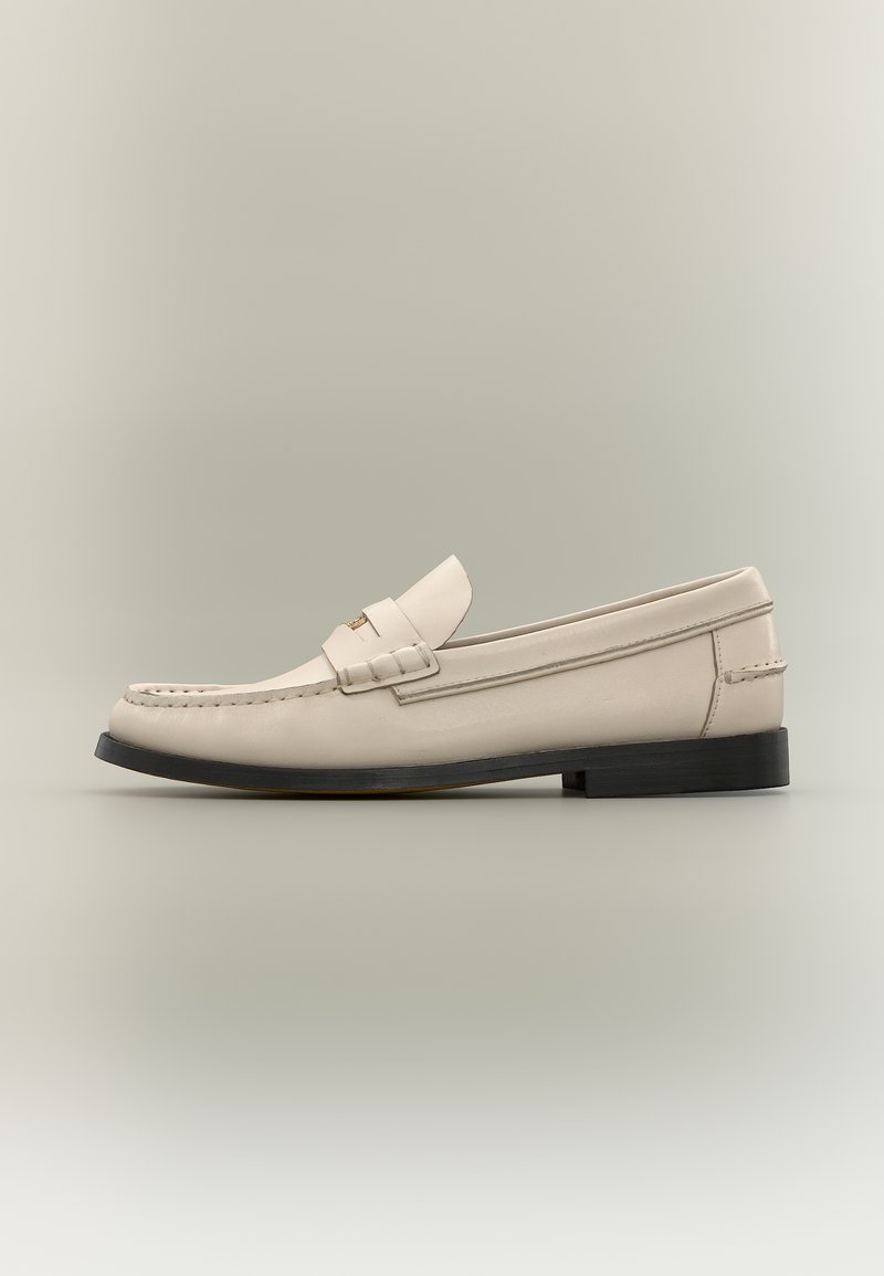 Μπεζ δερμάτινο μοκασίνι ανδρικού τύπου με μαύρη σόλα και διακοσμητική λωρίδα, σε ανοιχτό γκρι φόντο.