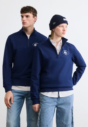 Twee modellen dragen marineblauwe Lacoste sweatshirts met een kwart rits, de een met lichte spijkerbroek en de ander met cargobroek en een marineblauwe muts.