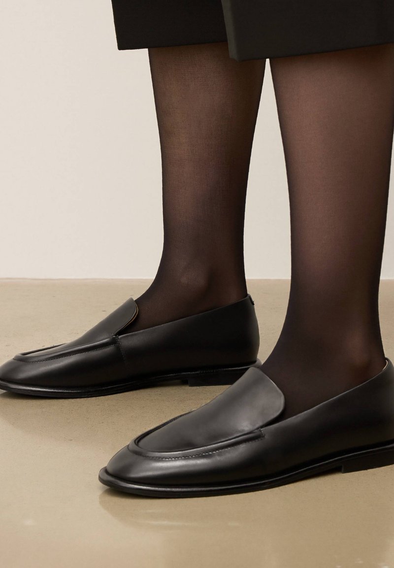 Mocassins en cuir noir avec une finition lisse, bout rond et détail de couture minimal, portés avec des collants noirs transparents. Semelle plate.