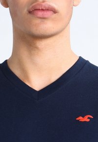 Närbild av en persons nedre ansikte och nacke som bär en marinblå V-ringad tröja med en liten röd fågel-logga på bröstet.