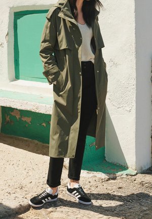 Manteau long vert olive avec poches à rabat, porté sur un pull beige et un pantalon noir taille haute. Des baskets noires et blanches complètent la tenue.