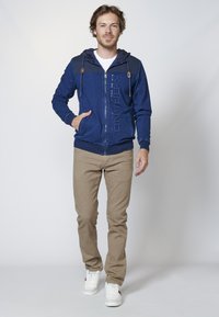 Navy blue zip-up hoodie with a textured hood, logo detail, and front pockets, paired with beige pants and white sneakers.