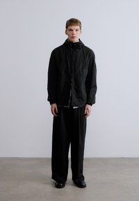 Must kapuutsiga jope üle ruudulise särgi, kombineeritud lahtiste mustade pükste ja mustade kingadega. Sile tekstuur, minimalistlik disain.