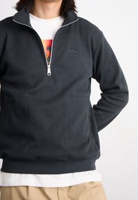 Mörkgrå zip-up sweatshirt med ribbat mönster, med en ståkrage och en diskret grön logotyp på bröstet. Vit undertröja synlig nedanför.