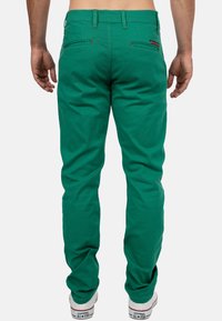 Pantaloni di cotone verde con un taglio slim, dotati di due tasche posteriori e dettagli di cucitura rossa. Indossati con sneakers bianche su uno sfondo neutro.