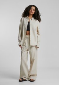 Beige oversized knäppskjorta och löst sittande byxor i lätt tyg. Svart croppad topp under. Platta sandaler med medelhög sula.