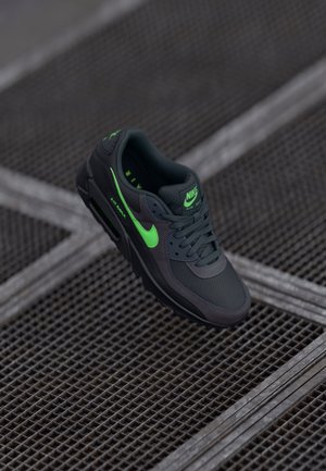 Baskets Nike Air Max noires avec un swoosh et logo vert vif, placées sur une surface en grille métallique.