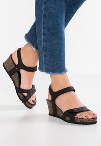 Sandali con tacco a cuneo neri con cinturini regolabili, base a effetto sughero, design open-toe e suola in gomma morbida. Jeans in denim sfilacciati visibili.