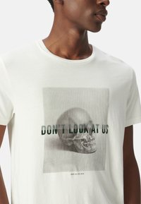 Camiseta de algodón blanca con un gráfico de una calavera y el texto verde "NO NOS MIRES" superpuesto. Mangas cortas y escote redondo.