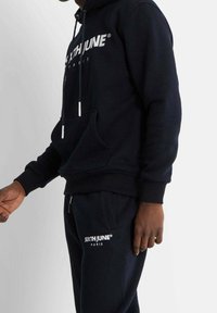 Sweat à capuche en coton bleu marine avec logo blanc, cordons de serrage et poche ventrale, assorti à un jogging assorti avec logo sur la jambe.