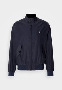 Veste bleu marine zippée avec col haut, poches à rabat boutonnées, poignets et bas côtelés, et petit logo poitrine côté gauche.