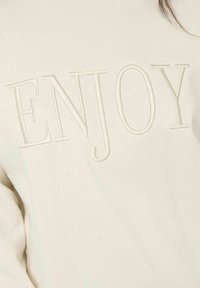 Tissu couleur crème avec de grandes lettres brodées en relief écrivant "ENJOY" sur la zone de la poitrine.