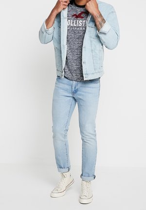Hellblaues Denim-Jacket über einem grauen Shirt mit Logo, kombiniert mit hellblauen Jeans und cremefarbenen Sneakers. Einfaches, lässiges Outfit.