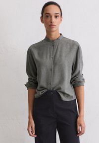 Green button-up shirt, textured fabric, high neckline, rolled sleeves. Paired with navy trousers featuring a front button closure.