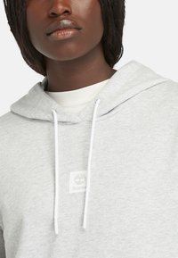 Svijetlo siva kapuljača napravljena od mekog pamuka, s bijelim užetom, rebrastim manžetama i ušivenim Timberland logom.