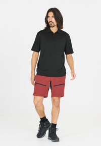 Svart pikétröja med korta ärmar, ihop med röda shorts med dragkedjefickor. Modellen har på sig svarta vandringsskor och strumpor.