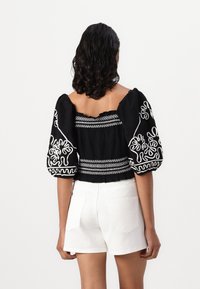 Svart broderte bluse med puffermer, med hvite intrikate mønstre og smocket rygg. Kombinert med hvite shorts, minimalistisk design.
