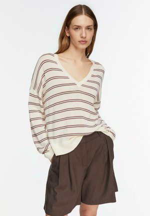 Kobieta w kremowym swetrze z dekoltem w serek z cienkimi bordowymi paskami i luźnych brązowych plisowanych szortach, pozująca z rękami w kieszeniach.