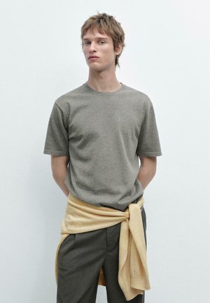 Jonge man met lichtbruin haar die een grijs T-shirt met korte mouwen, een donkere broek en een gele trui om zijn middel geknoopt draagt.