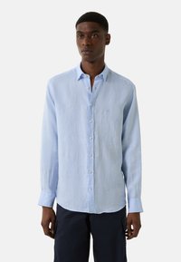 Lichtblauwe blouse met lange mouwen van linnen, voorzien van een button-down kraag, witte knopen en een geborduurd logo op de borst. Zachte textuur en een ontspannen pasvorm.