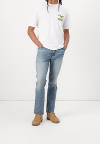 Valkoinen puuvillapaita graafisella printillä, siniset farkut ja vaaleanruskeat mokkakengät. Suora lahje, rento malli, kengissä ei näkyviä yksityiskohtia.