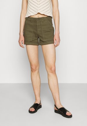 Person iført olivengrønne shorts med op rullede kanter, en cropped beige strik top og sorte åbne sandaler, der står på et lysegråt gulv.